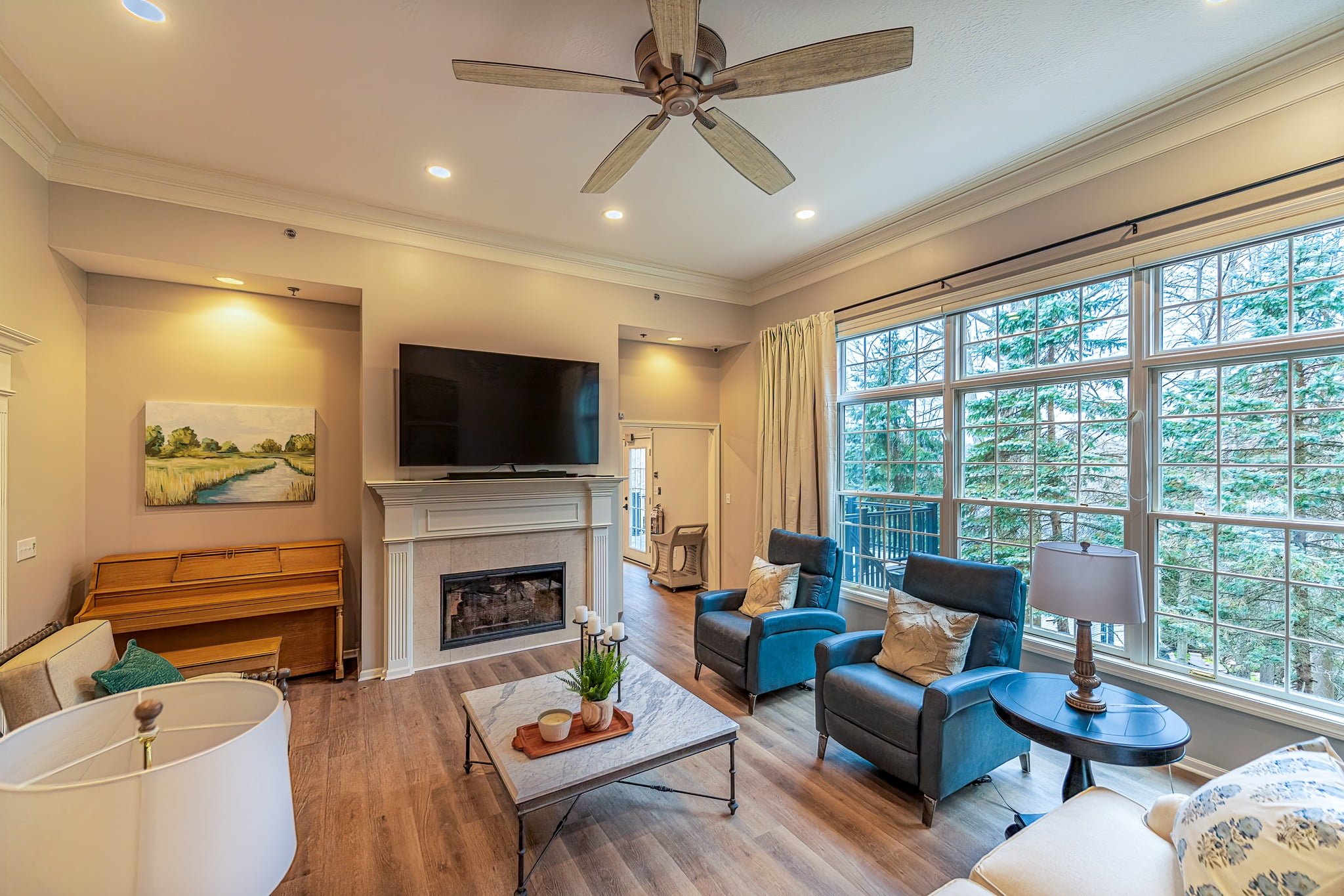 Common living and activity area featuring a piano, glassed fireplace, big TV, open spaces, comfortable seating, and panoramic windows overlooking a wooded area at Camella Living.