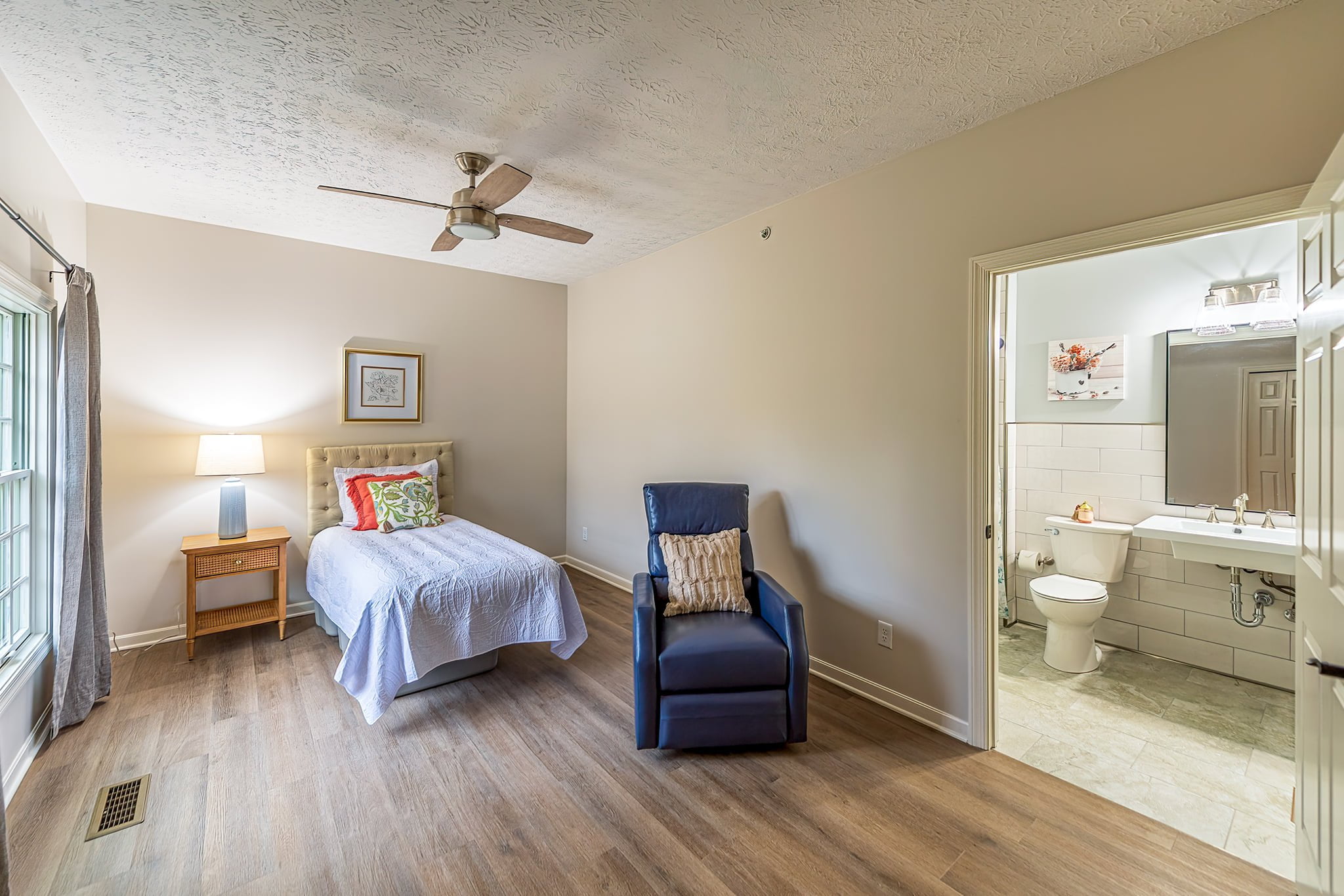 Spacious and well-lit bedroom with a bed, cushions, adjustable recliner, and an accessible bathroom compliant with memory care guidelines at Camella Living.
